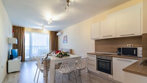 A modern kitchen with white cabinets, a microwave, and a stove. There is a dining area with a table and chairs. - Varna South Bay Apart Complex (Varna)