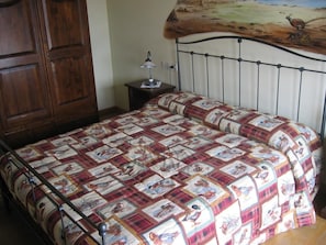 A bedroom with a bed covered in a patterned quilt, a wooden wardrobe, a bedside table with a lamp, and a wall painting. - La Valle (Giavera del Montello)