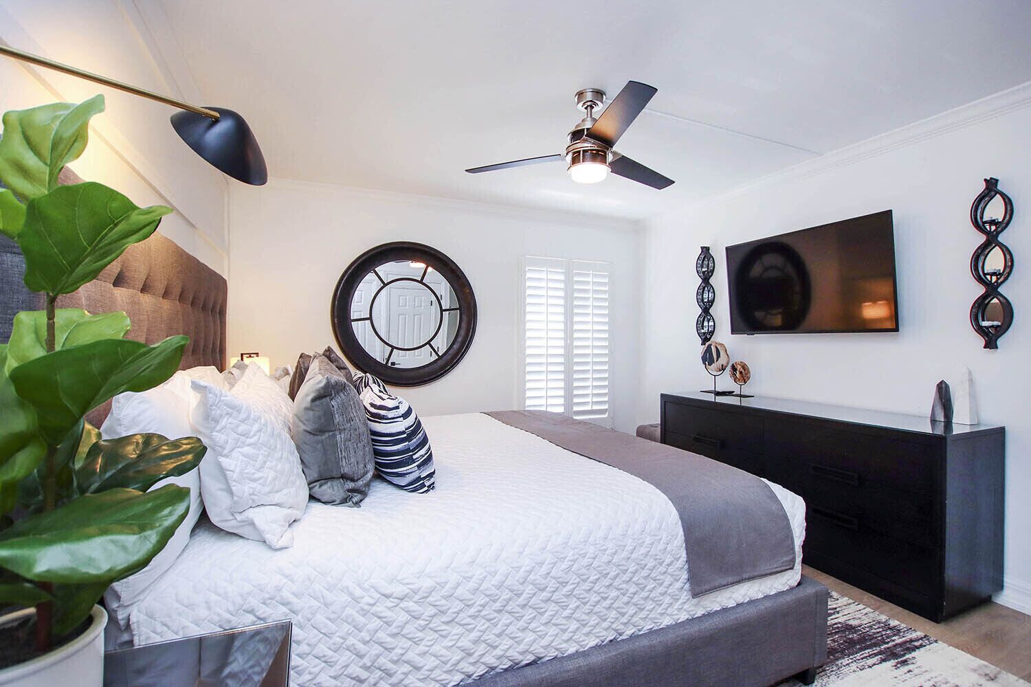 A modern bedroom with a bed, bedside table, TV, ceiling fan, and a large window with blinds.
