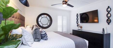 A modern bedroom with a bed, bedside table, TV, ceiling fan, and a large window with blinds.