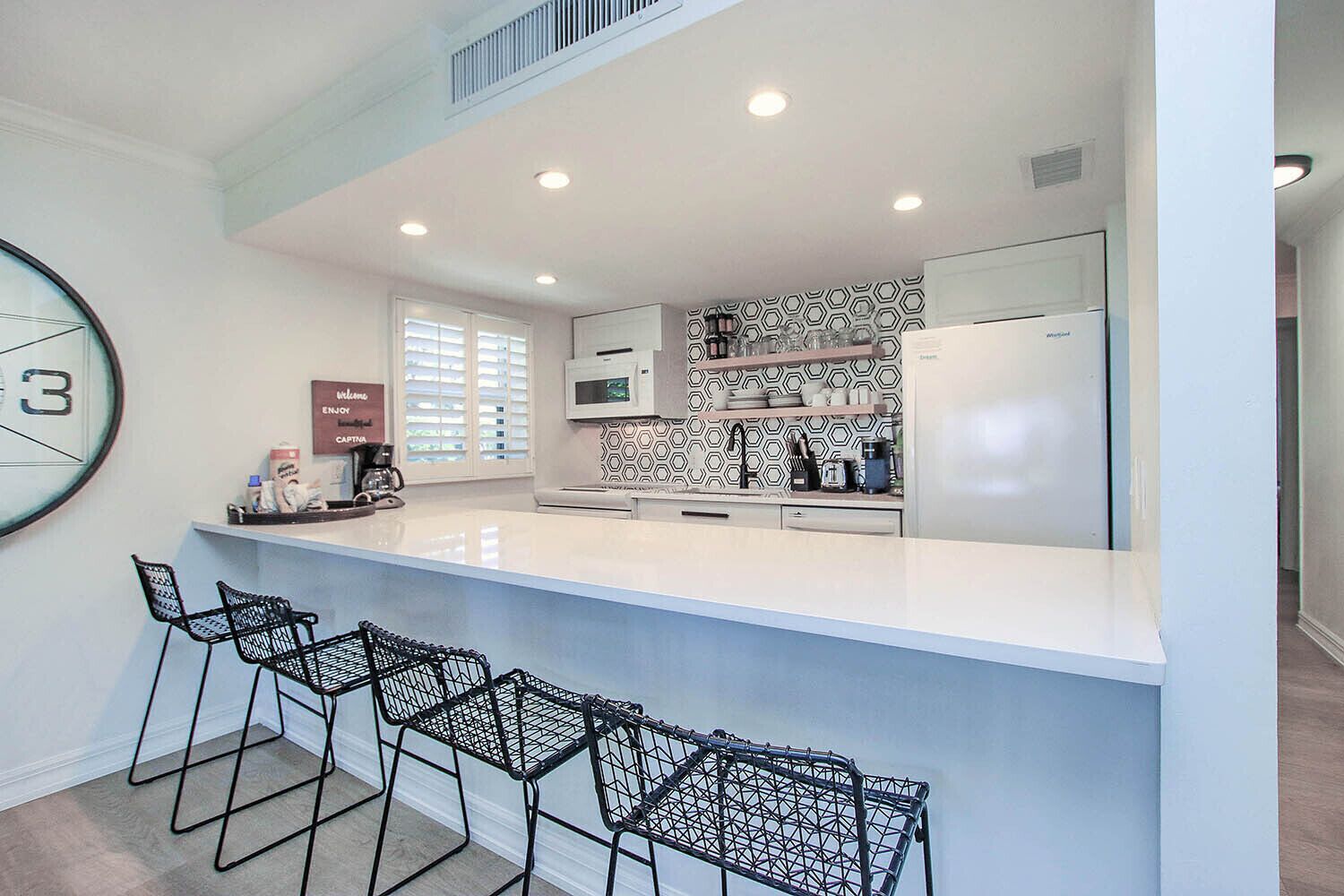 A modern kitchen with a white countertop, black bar stools, a microwave, and a refrigerator.