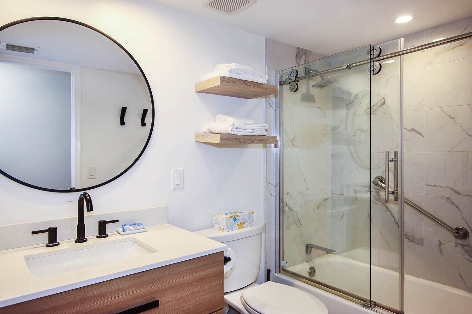 A modern bathroom with a round mirror, a sink with two faucets, a toilet, and a glass-enclosed shower with a handheld showerhead.