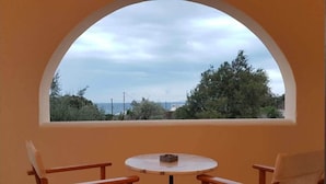 A small balcony with two wooden chairs and a table, overlooking a coastal landscape with trees and a view of the sea. - Dagris Villa Studios (Santorini)