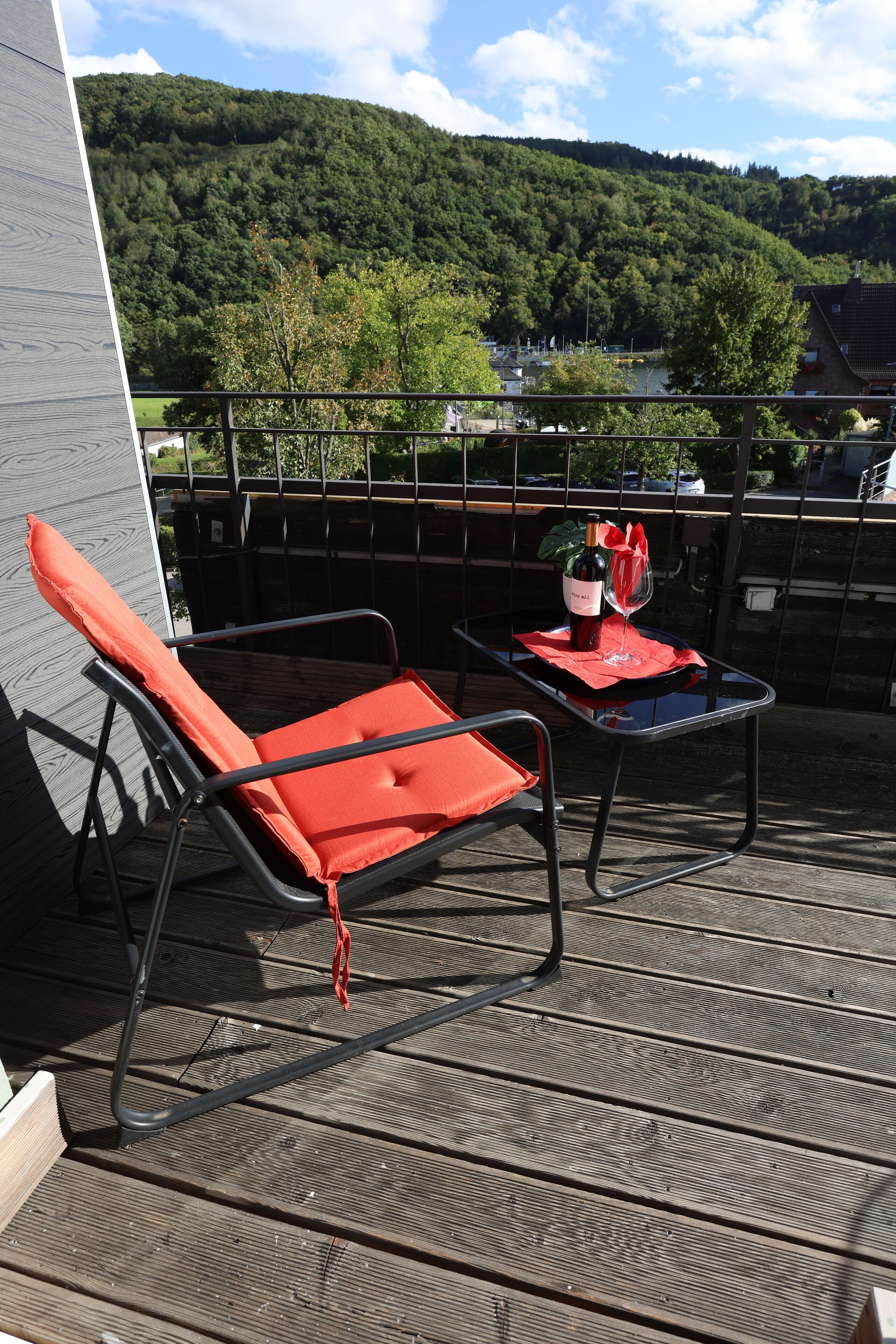 A balcony with a red lounge chair, a small table with a bottle and glass, and a view of a forested hill.