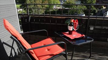 A balcony with a red lounge chair, a small table with a bottle and glass, and a view of a forested hill.