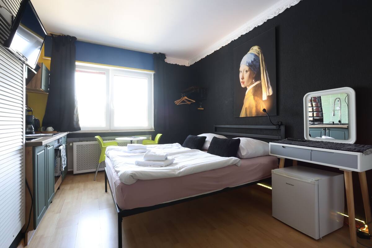 Kompakti hotellihuone, jossa on sänky, pieni jääkaappi, työpöytä tietokoneella, ikkuna verhoilla ja taulu seinällä.