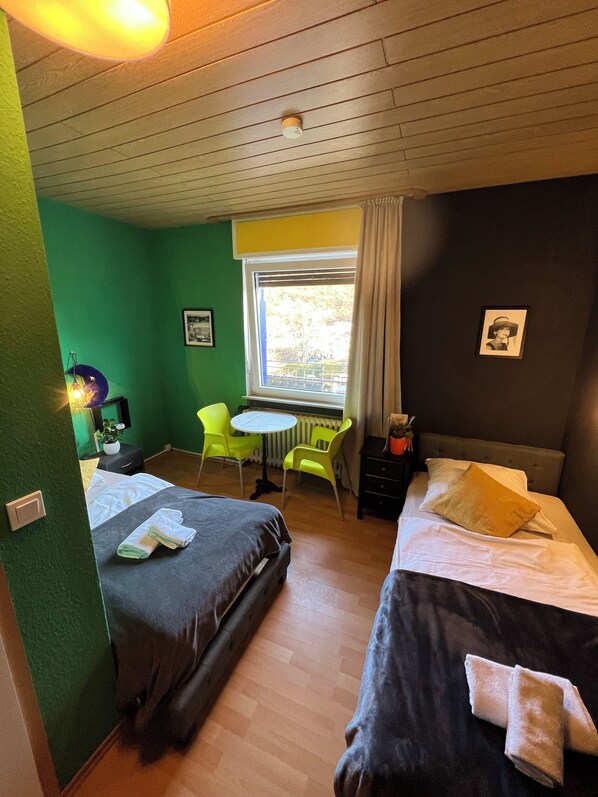 Habitación con dos camas, un escritorio y una silla. Se observa una ventana con cortinas, un radiador y una pequeña mesa con una planta.