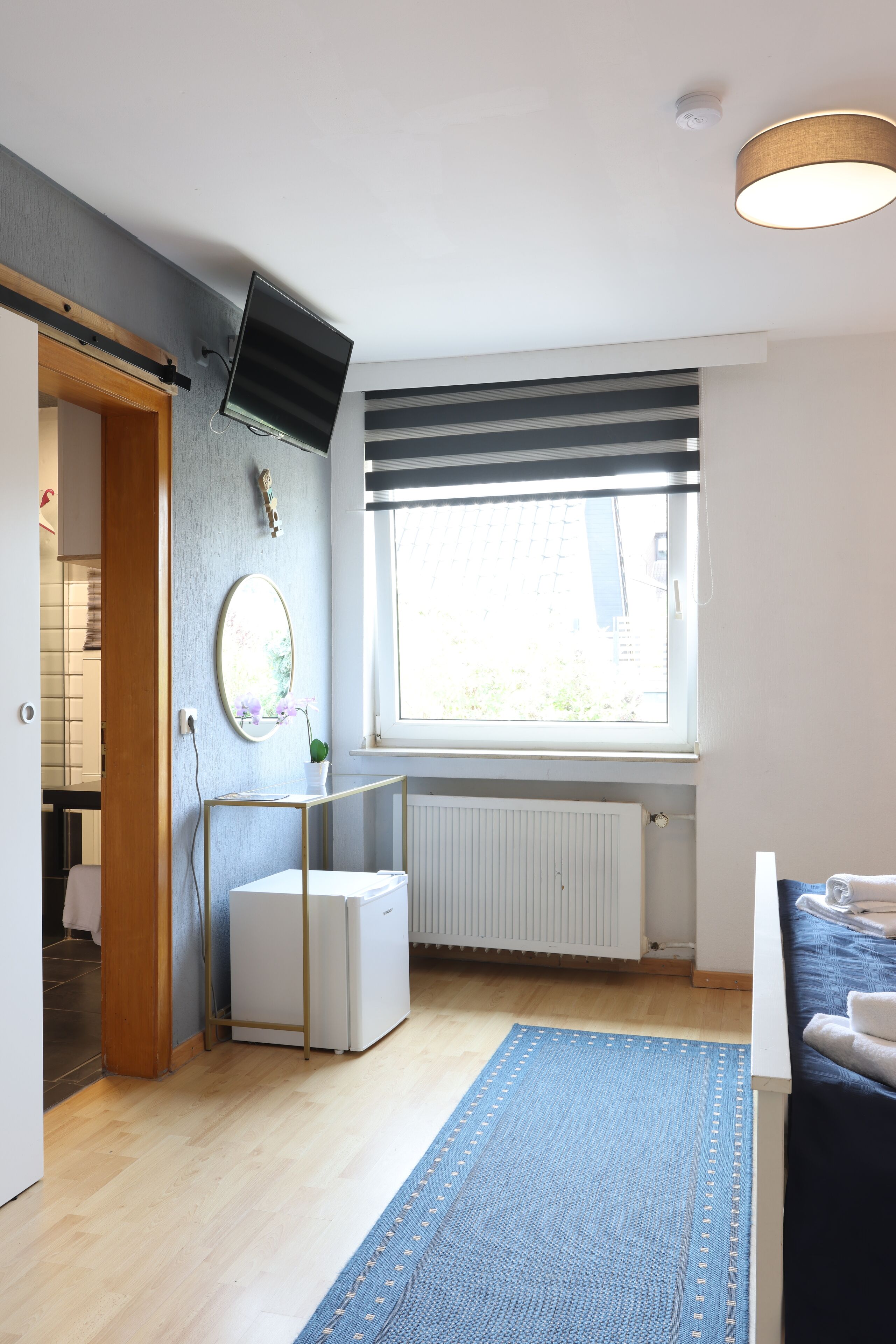 Una habitación moderna con un ventanal grande, un televisor de pantalla plana montado en la pared, un radiador debajo de la ventana y una alfombra azul en el suelo.