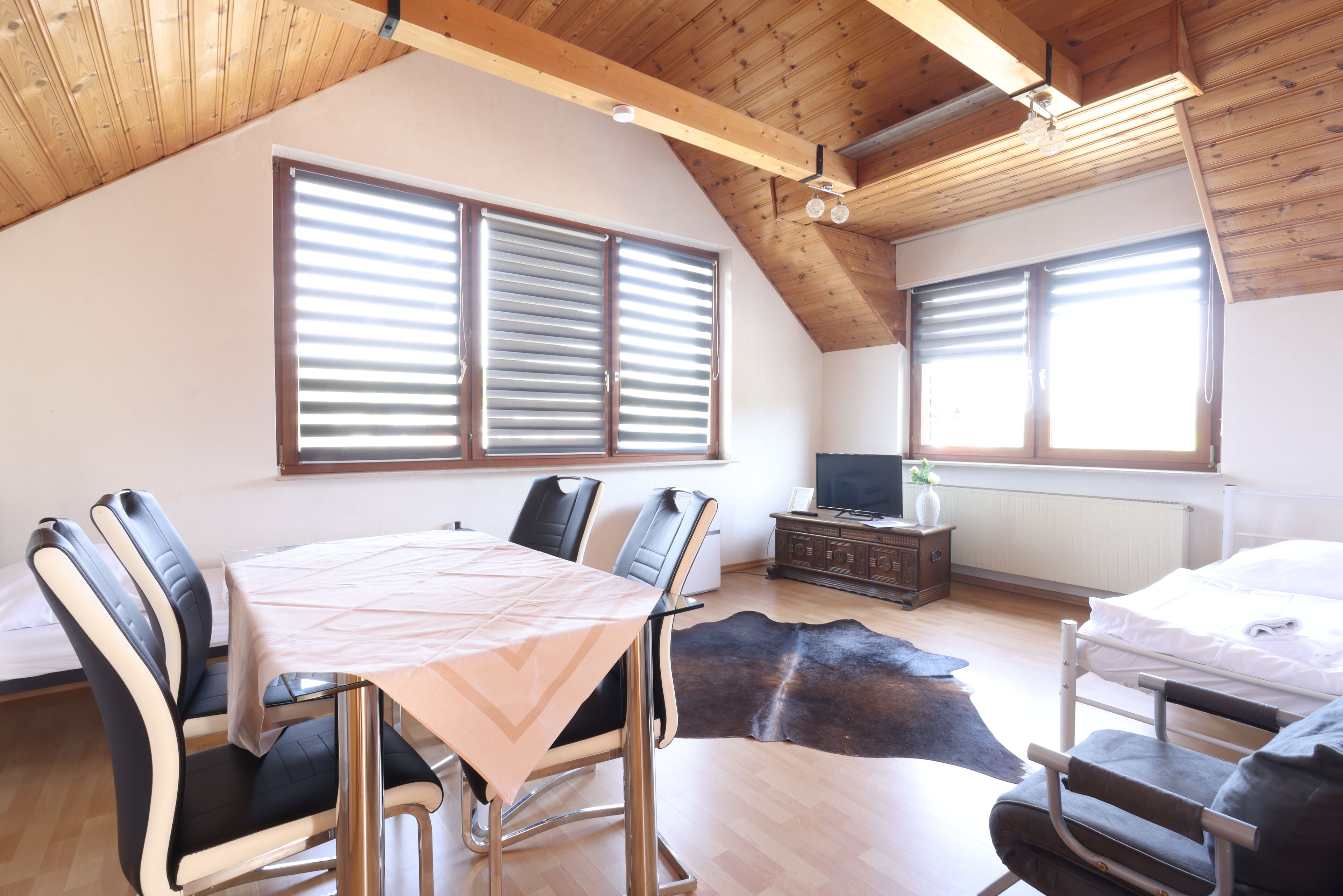 A room with a dining table, chairs, a bed, a TV, and large windows with blinds.