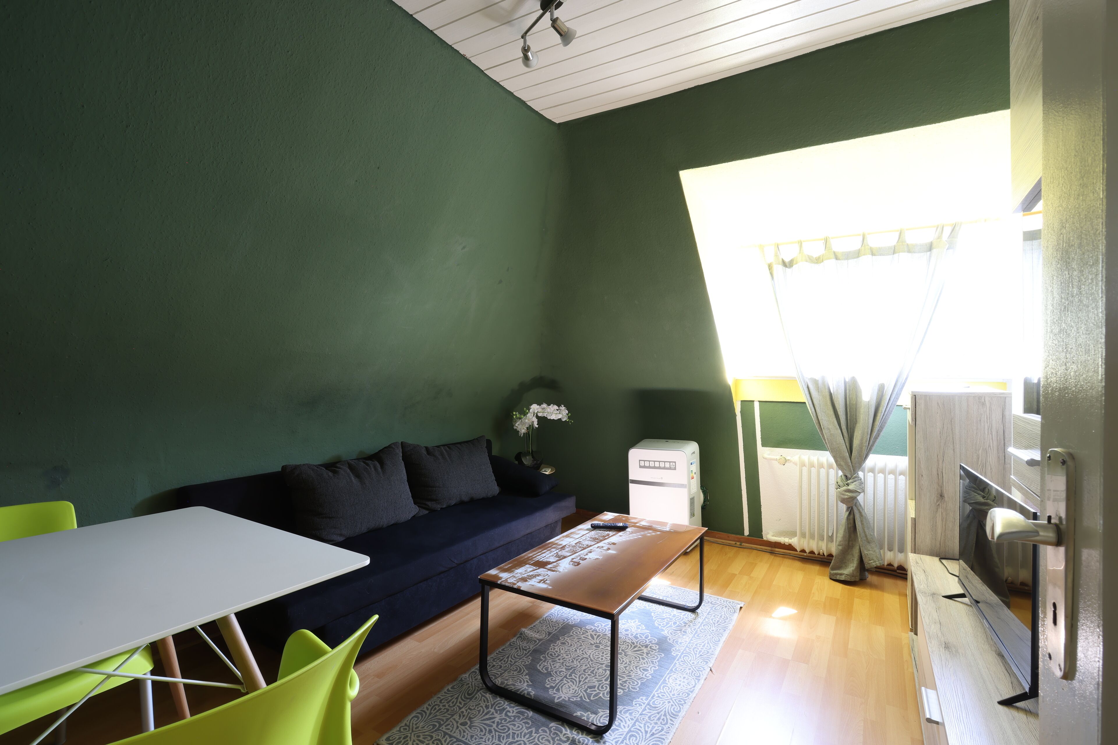 Una habitación con pared verde, una mesa blanca con sillas, un sofá, un radiador y una ventana con cortinas.