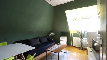 Una habitación con pared verde, una mesa blanca con sillas, un sofá, un radiador y una ventana con cortinas.