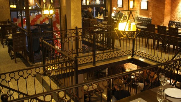 Un restaurant amb zona de bar, una escala i una sala de dinar amb taules i cadires.