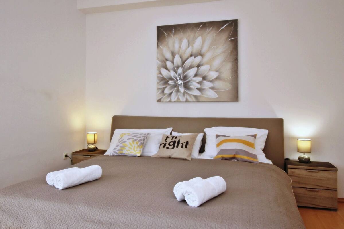 Una cama bien hecha con dos toallas, una obra de arte floral grande en la pared y dos lámparas de noche.