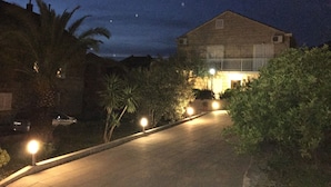 Vista noturna de um edifício com telhado inclinado, rodeado por vegetação e um caminho pavimentado.