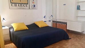 Een slaapkamer met een bed, twee gele kussens, een nachtkastje, een bureau, een stoel, een garderobe en een plank met een blauw object.