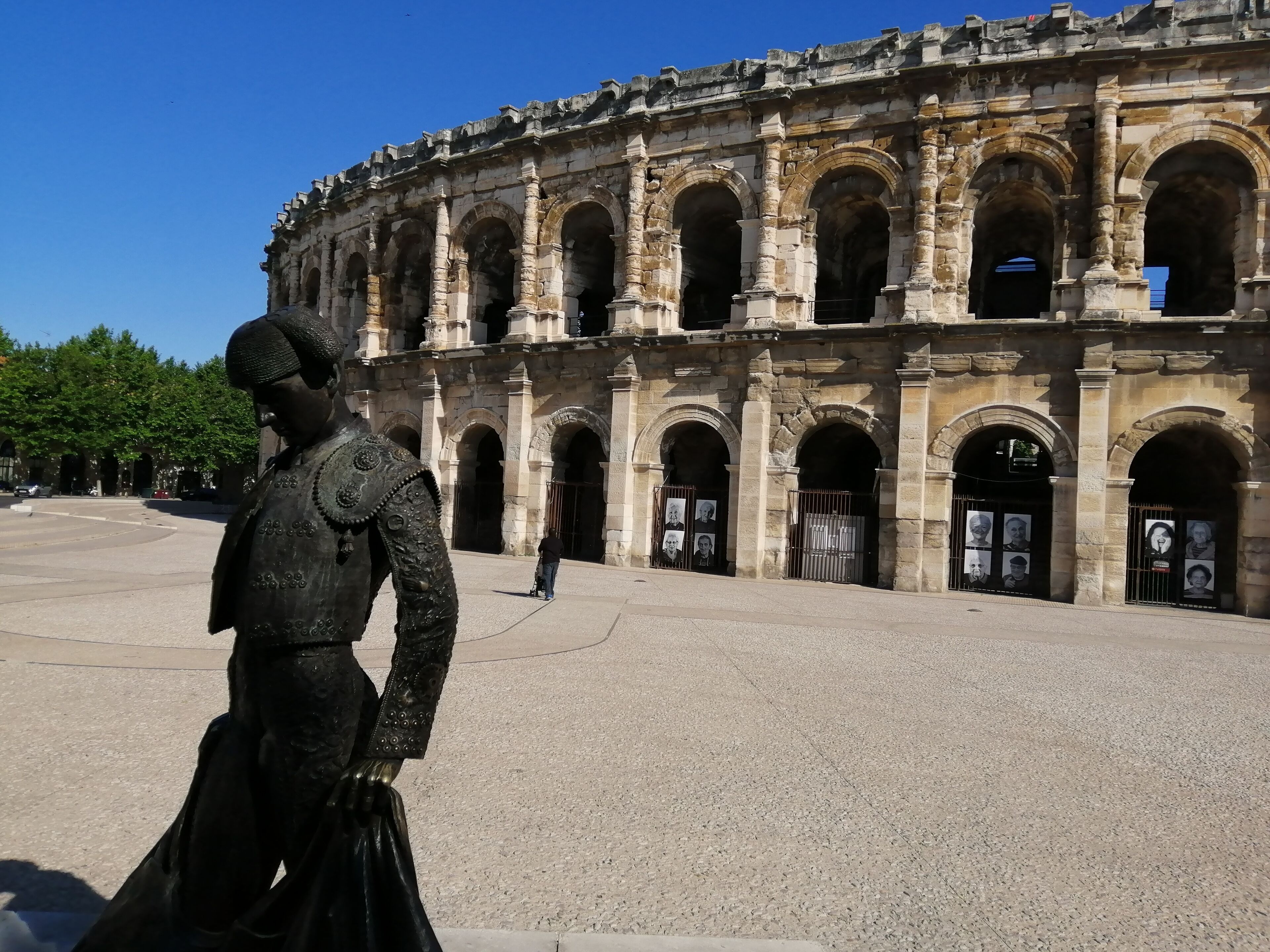A historical building with multiple arches and columns, a statue in the foreground, and a person standing in front.