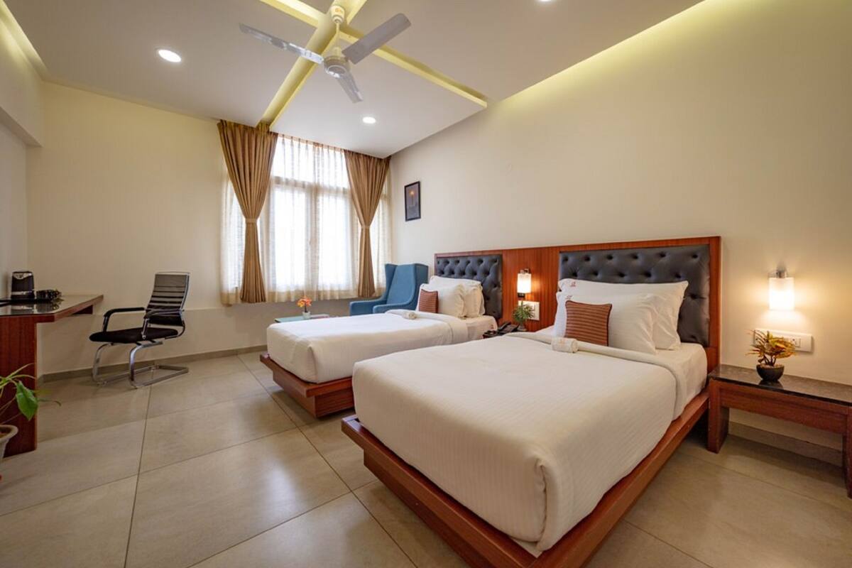 Une chambre d’hôtel avec deux lits, un bureau, une chaise, un ventilateur de plafond et une fenêtre avec des rideaux.
