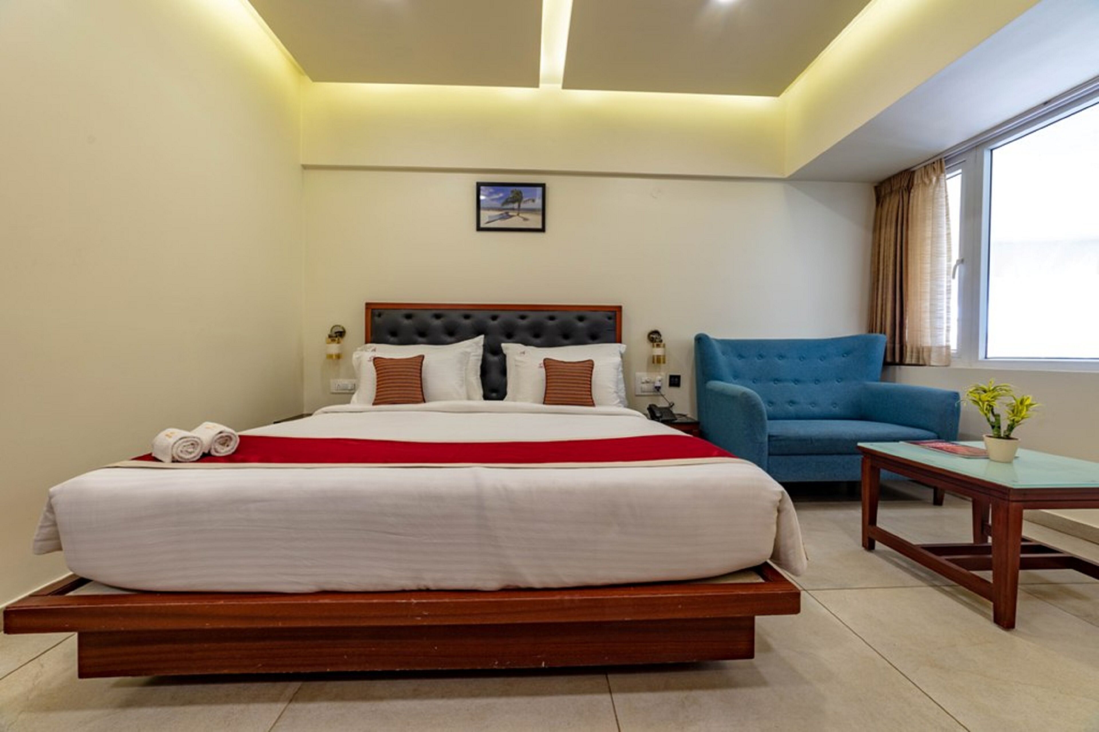 A hotel room with a large bed, a blue sofa, a small table with a plant, and a window with curtains.
