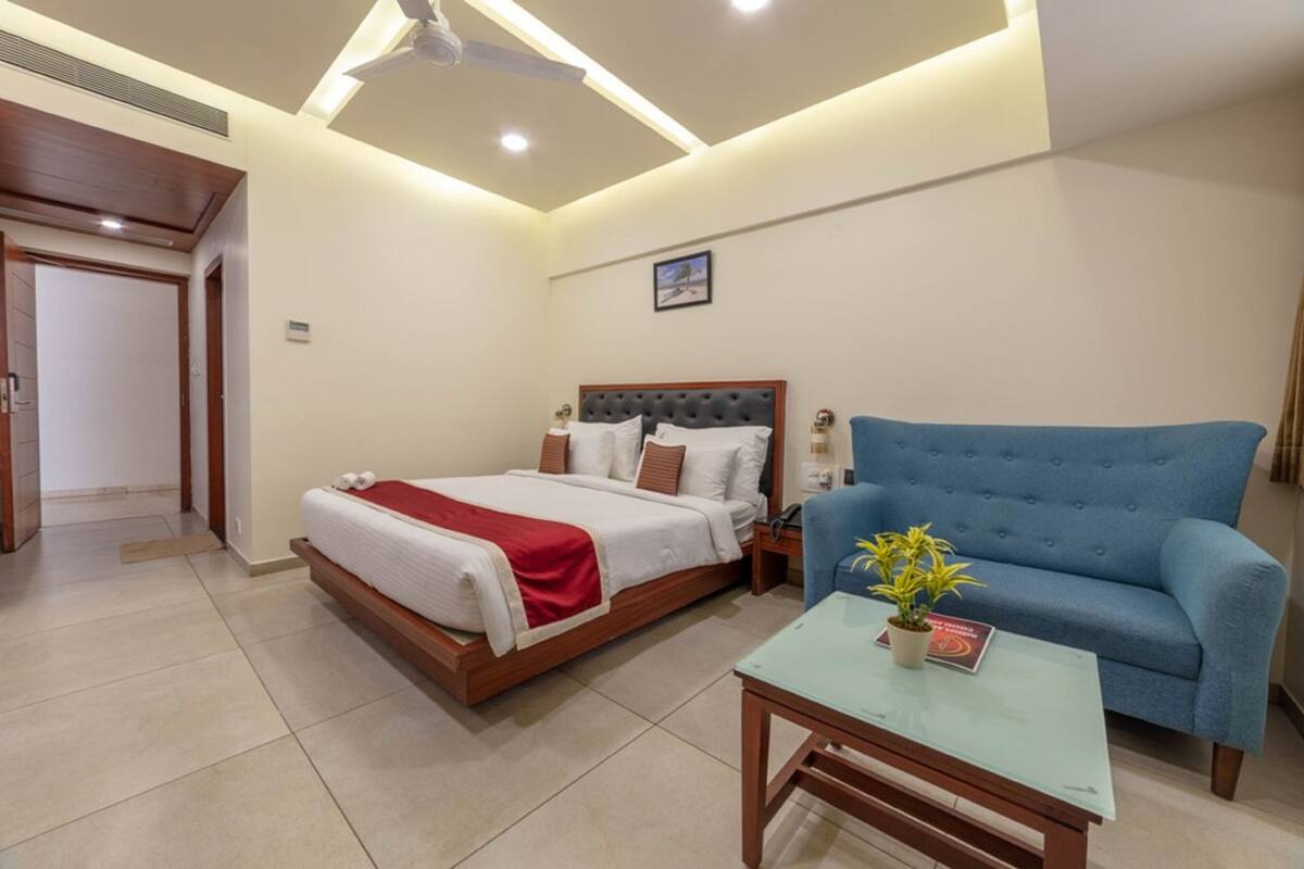Une chambre d’hôtel avec un lit, un canapé bleu, une petite table avec une plante et un ventilateur de plafond.