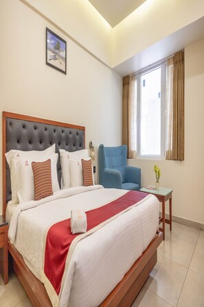 A hotel room with a large bed, a blue armchair, a small table, and a window with curtains.