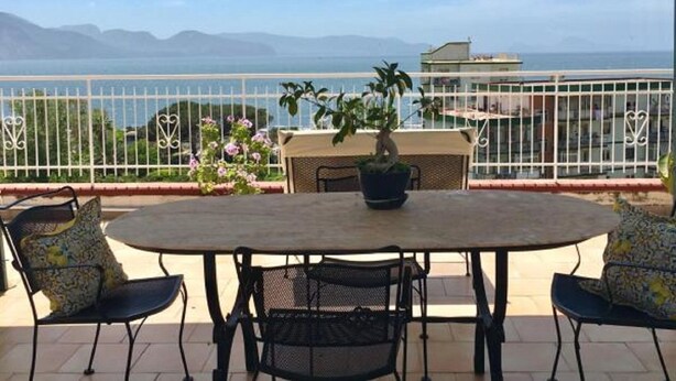 Ein Balkon mit Tisch und Stühlen, einem Blumentopf und Blick auf das Meer und die Berge.