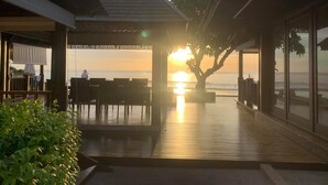 A wooden deck with a view of the sunset over the ocean, a covered seating area, and a large tree in the center.