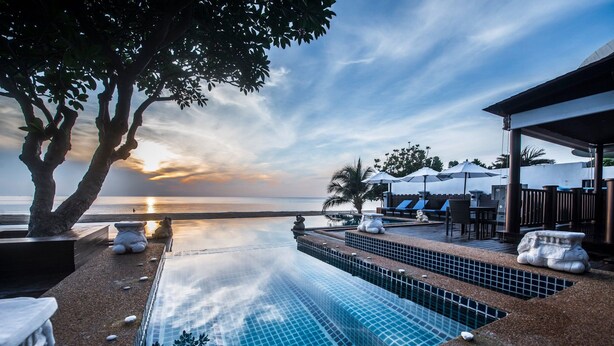 A infinity pool with a view of the sunset over the ocean, surrounded by stone seating and umbrellas.