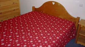 Una cama individual con una colcha roja estampada con flores, cabecera de madera con un corte en forma de corazón y una mesita de noche de madera con una lámpara.