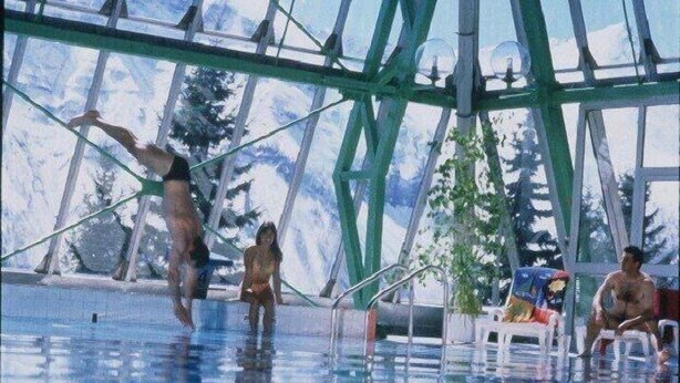 Una piscina con techo de cristal, rodeada de árboles y algunas personas.