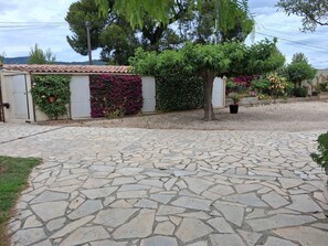 A stone-paved driveway leading to a building with a tiled roof, surrounded by lush greenery and flowering plants.