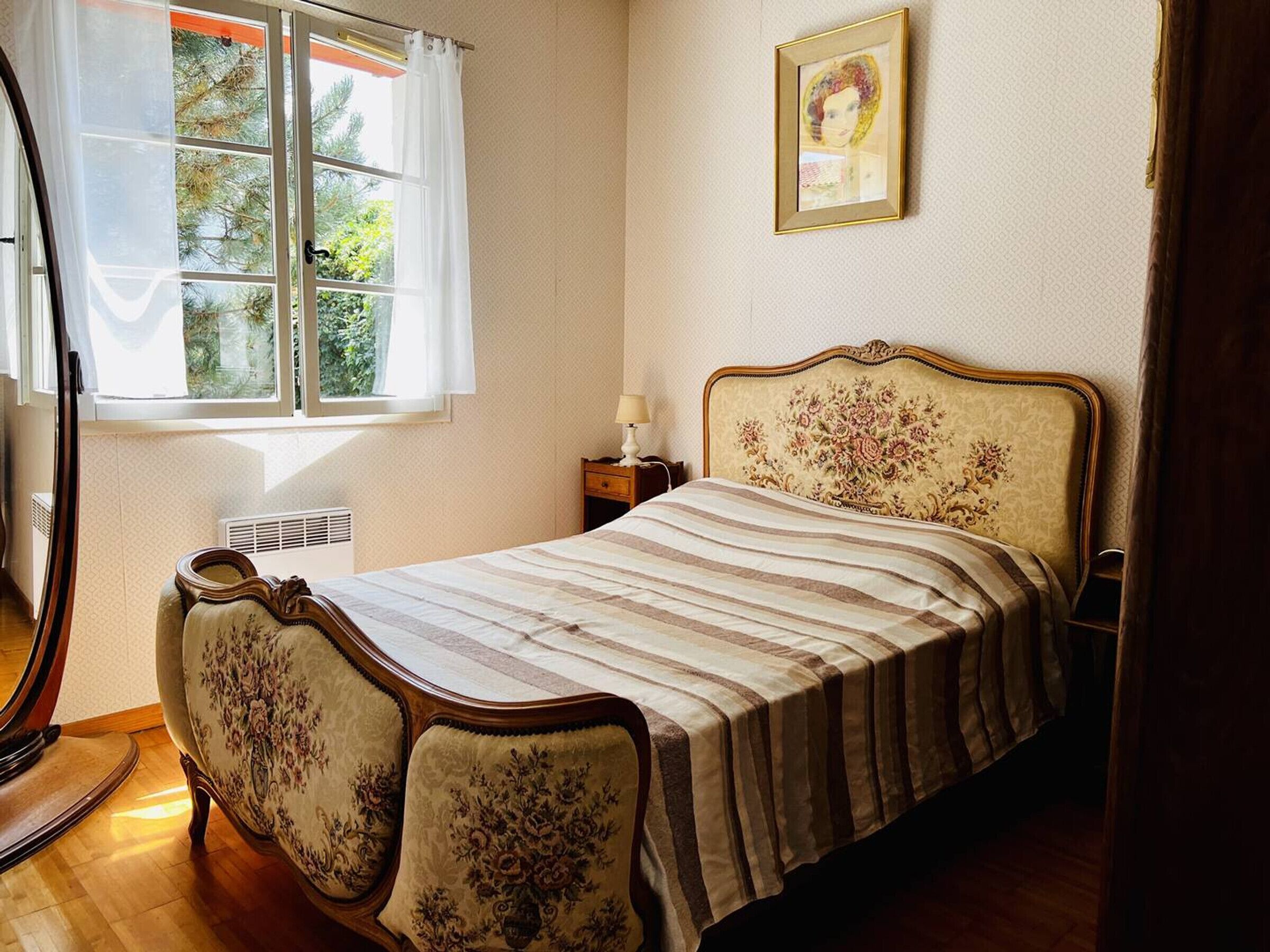 Une chambre à coucher avec un lit, une table de chevet, une lampe, un tableau encadré au mur et une fenêtre avec des rideaux.