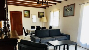 Una sala de estar con sofá, una mesa de centro y una zona de comedor con sillas y mesa. Hay cuadros en las paredes y una escalera de madera.