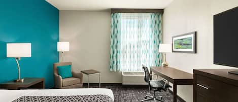 A hotel room with a bed, two bedside tables with lamps, a desk with a chair, a small table, a window with curtains, and a television on the wall.