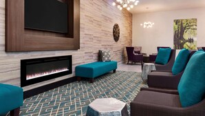 Un área de descanso moderna con un televisor de pantalla plana, chimenea, asientos en tonos aguamarina y marrón, alfombra estampada y un reloj fijado a la pared.
