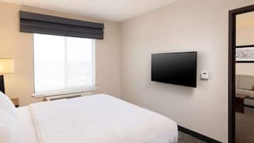 A hotel room with a bed, a flat-screen TV mounted on the wall, a window with a dark valance, and a door leading to another room.