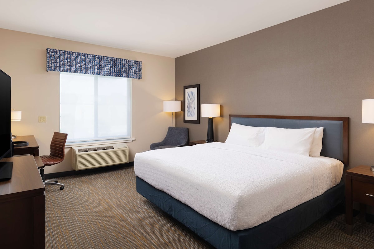 A hotel room with a large bed, a desk with a chair, a window with blinds, two bedside lamps, and a framed picture on the wall.
