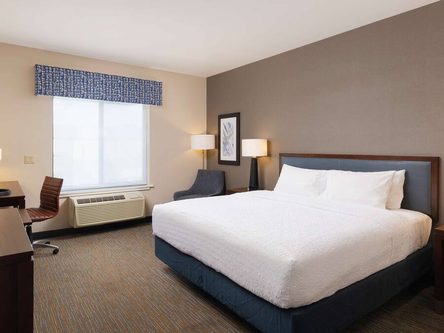 A hotel room with a large bed, a desk with a chair, a window with blinds, two bedside lamps, and a framed picture on the wall.