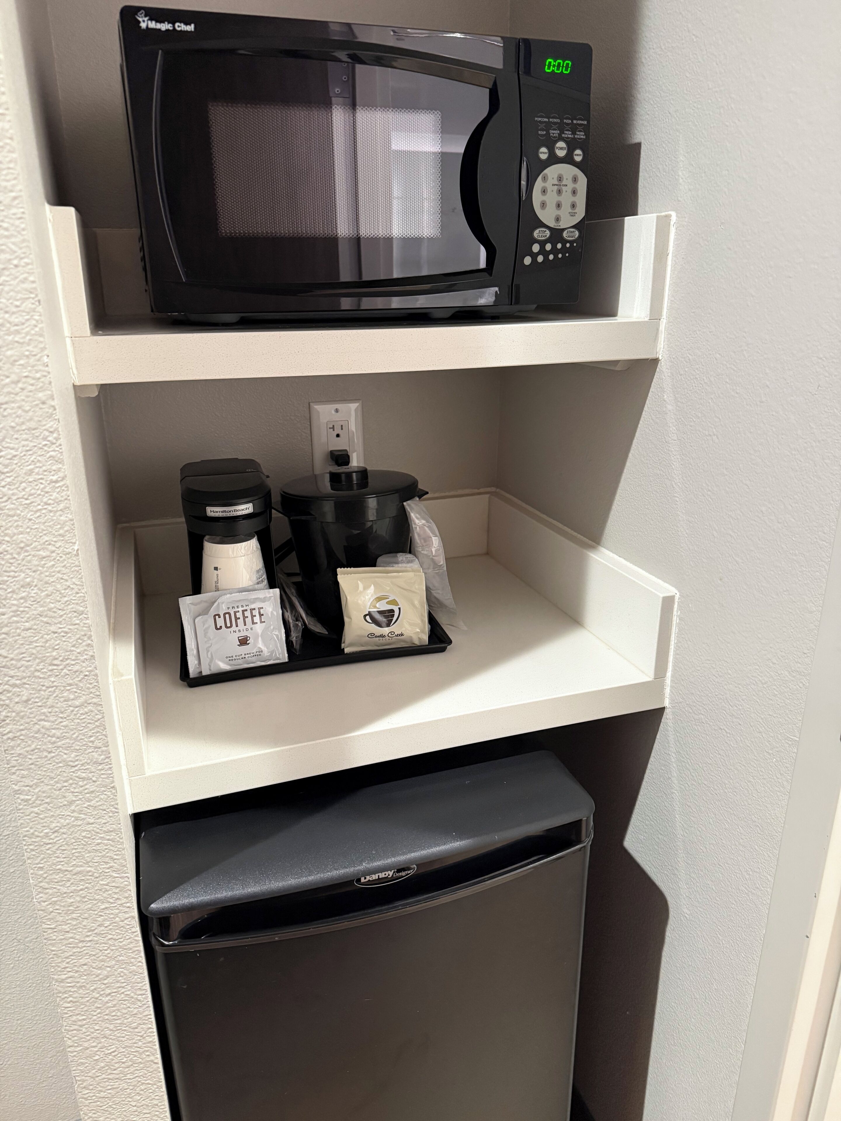 A kitchen area with a microwave on the upper shelf, a coffee maker and coffee pods on the lower shelf, and a refrigerator below.