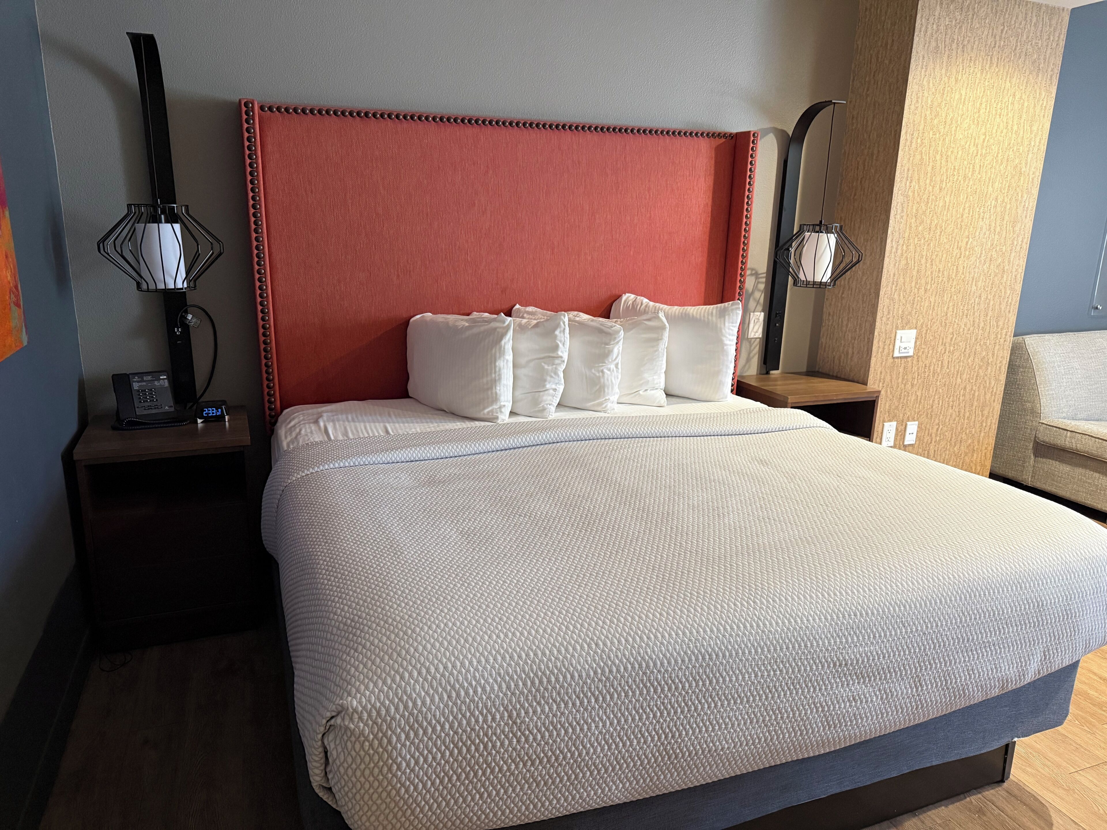 A hotel room with a large bed, a red upholstered headboard, white bedding, and a bedside table with a lamp.
