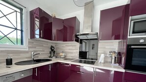 A modern kitchen with dark cabinets, a stainless steel range hood, a built-in oven, and a window providing natural light. - Cambo: Bright T2 with South Terrace, Wifi & Parking (Cambo-les-Bains)