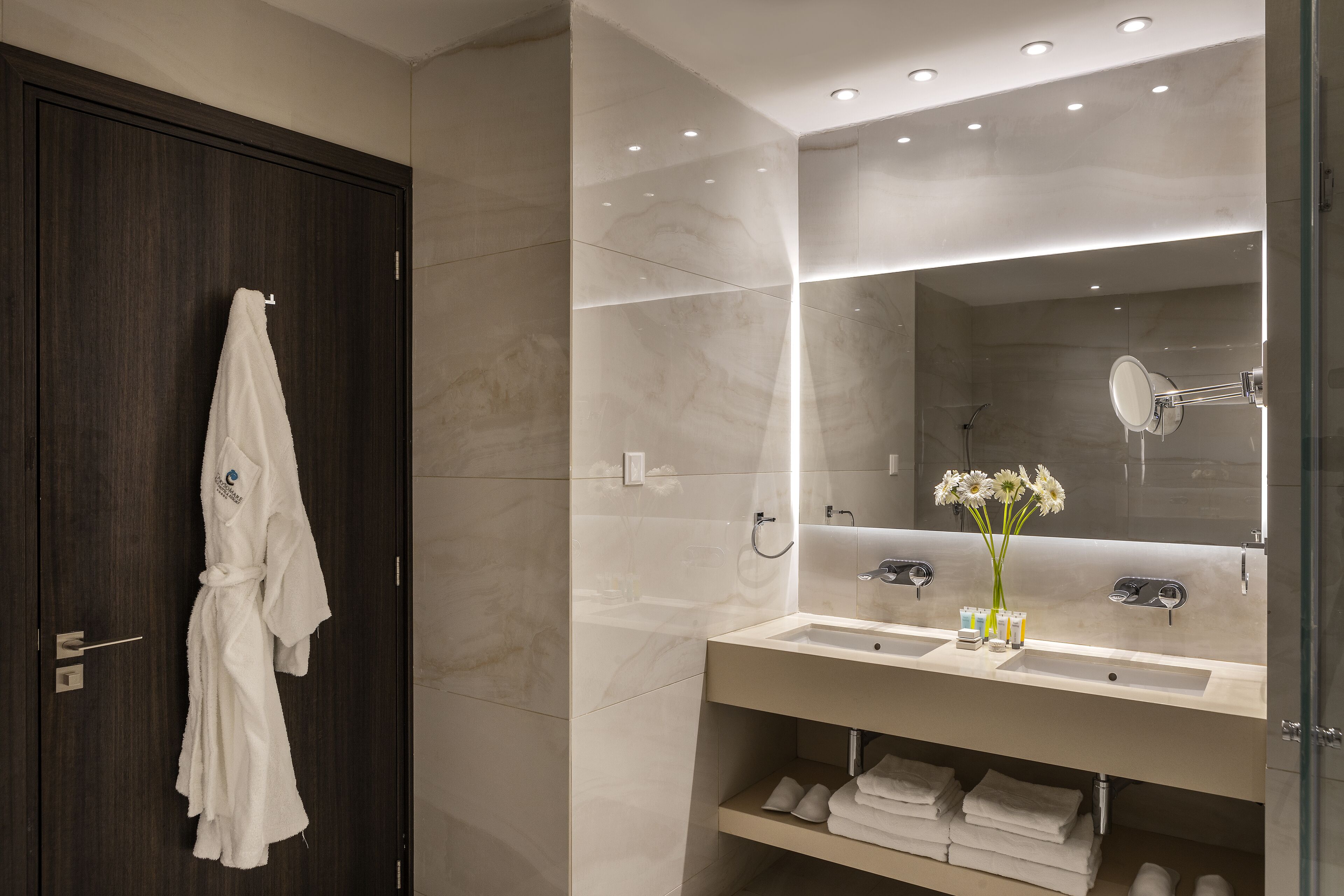 Une salle de bain moderne avec une porte en bois foncé, un grand miroir et un meuble-lavabo doté d’un évier et d’un espace de rangement pour les serviettes.