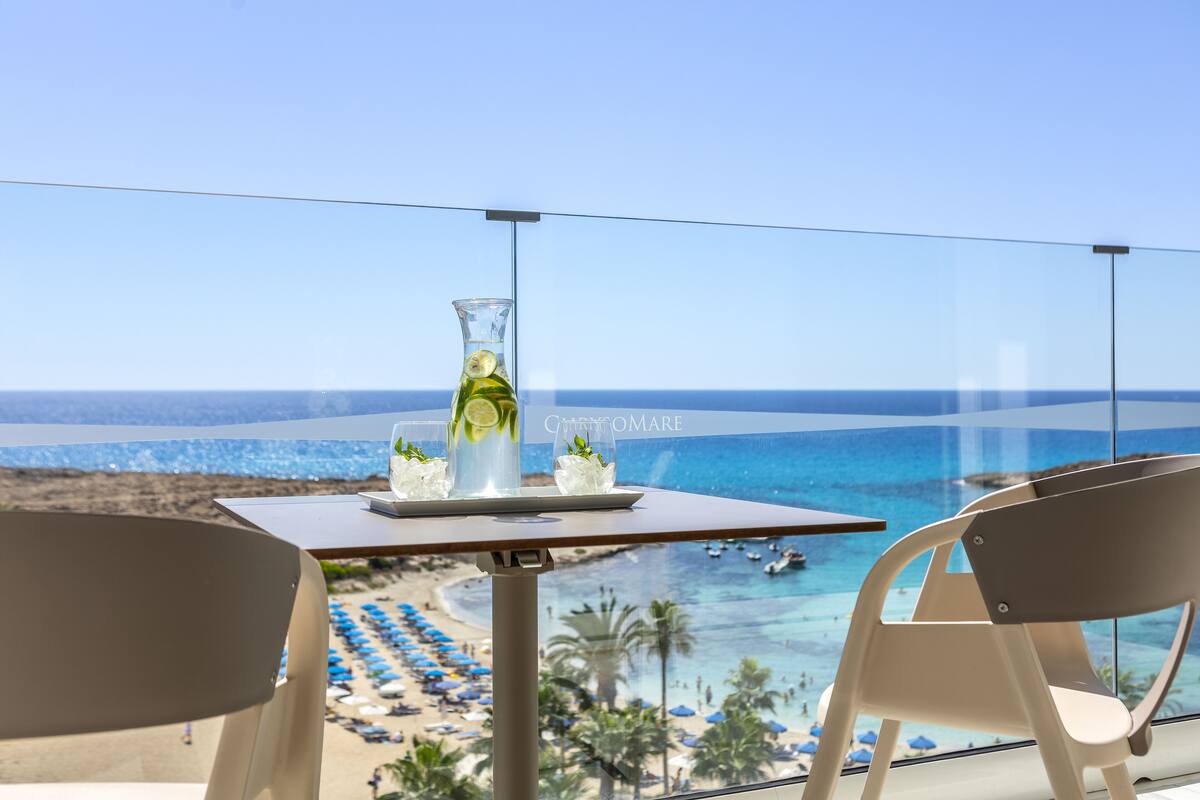 Un balcón con vista despejada a la playa, una mesa con un florero y flores, y dos sillas.