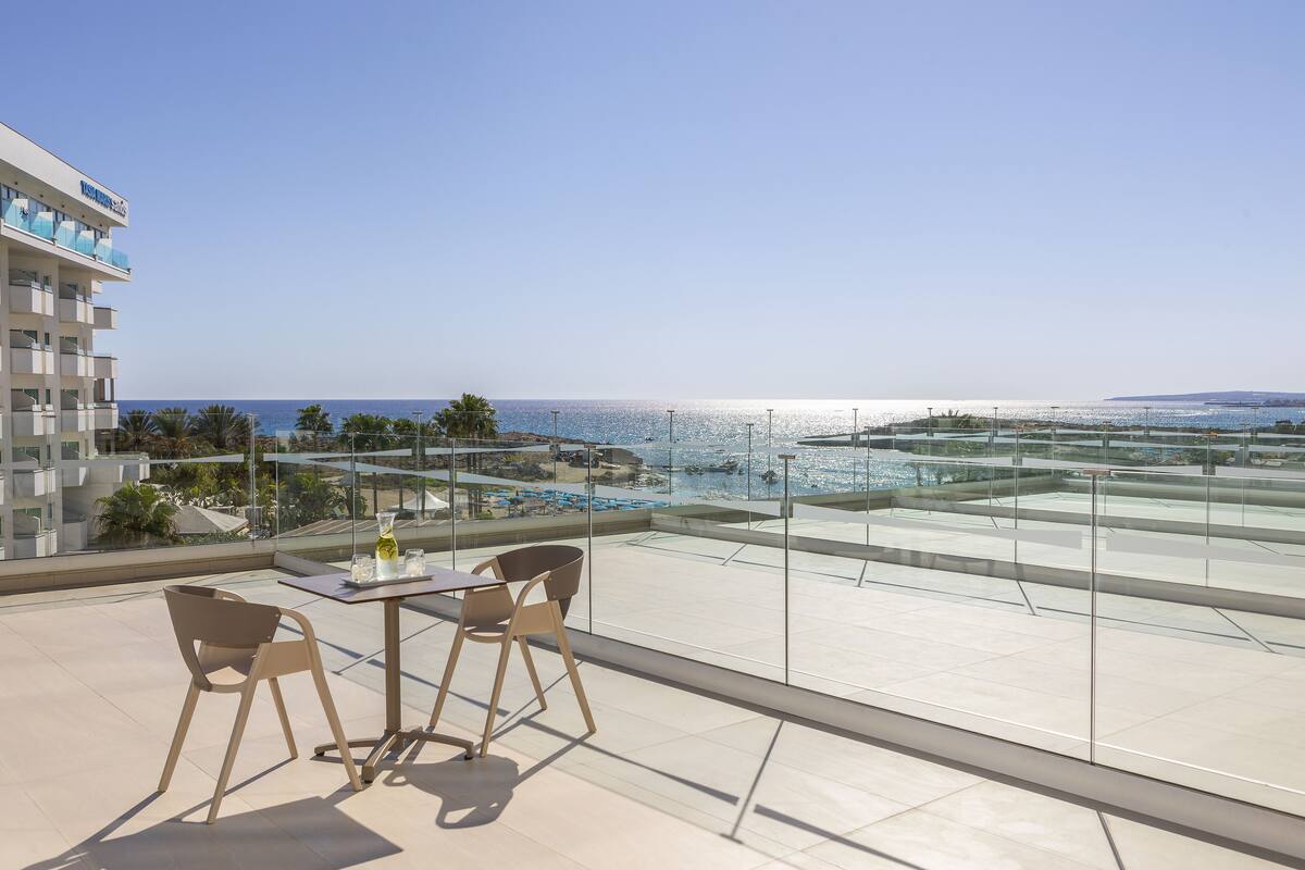 Una terraza en la azotea con barandilla de cristal, una mesa con sillas y vistas al mar y a la costa distante.
