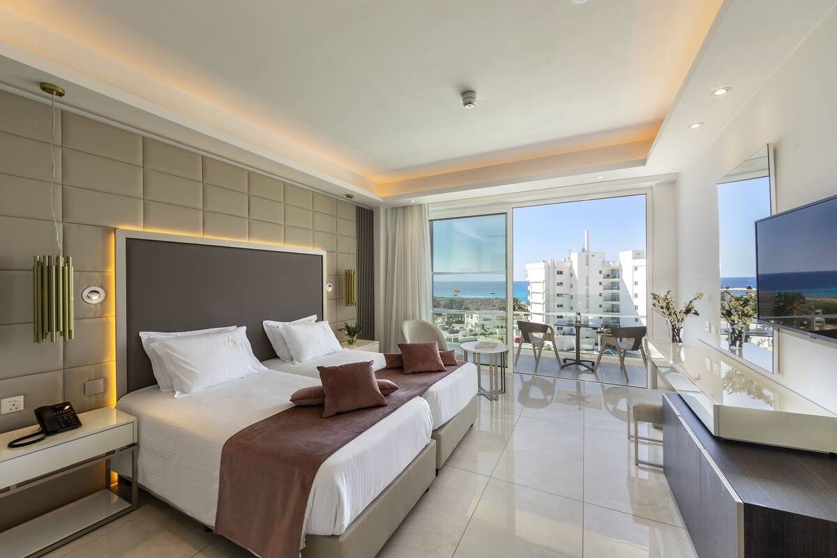 Una habitación de hotel moderna con una cama grande, mesitas de noche, un televisor y un balcón con vistas a la ciudad.