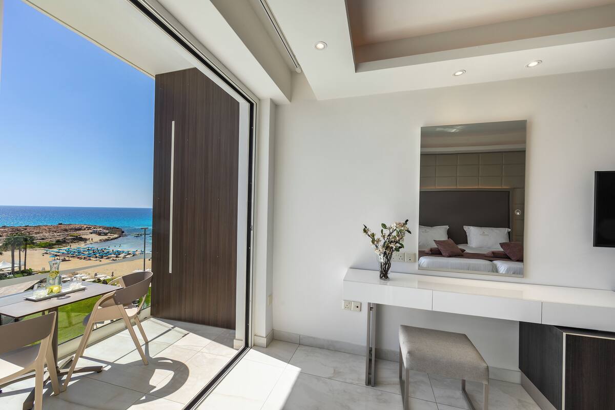 Una habitación moderna con balcón y vistas a la playa, zona de comedor y un espejo grande que refleja la cama.
