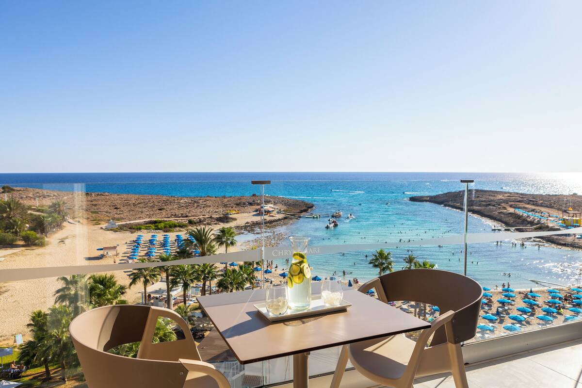 Un balcón con una mesa, sillas y una bebida. Se ve una playa, sombrillas y el mar.