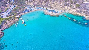 Luftaufnahme einer Küstenregion mit Strand, klarem, blauem Wasser und einem Parkplatz mit vielen Autos.