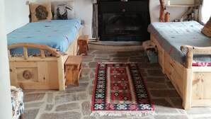 A rustic room with two wooden beds, a stone floor, a fireplace, and a stone wall with a painting.