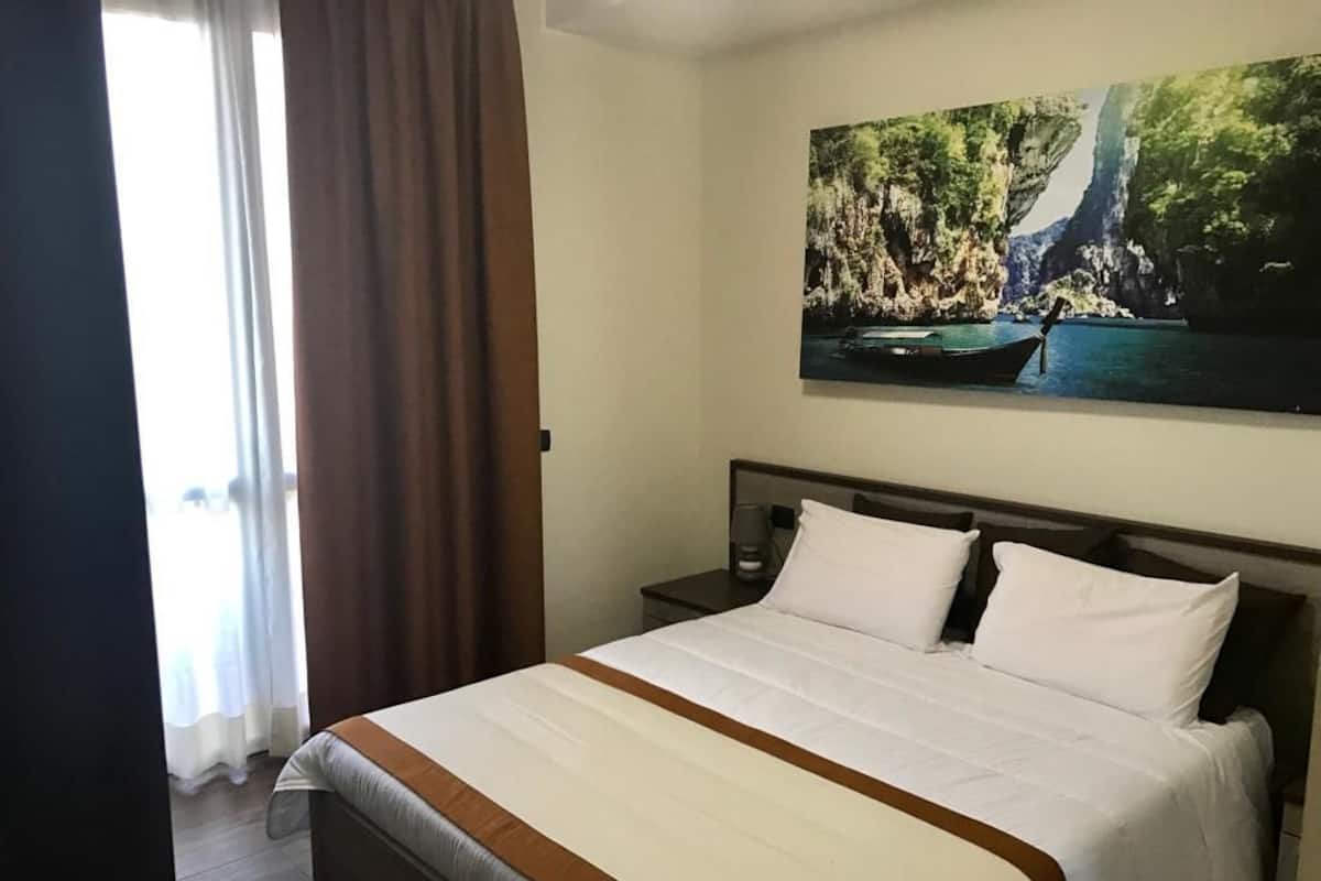 A hotel room with a large bed, a nightstand, a painting of a waterfall, and a window with curtains.