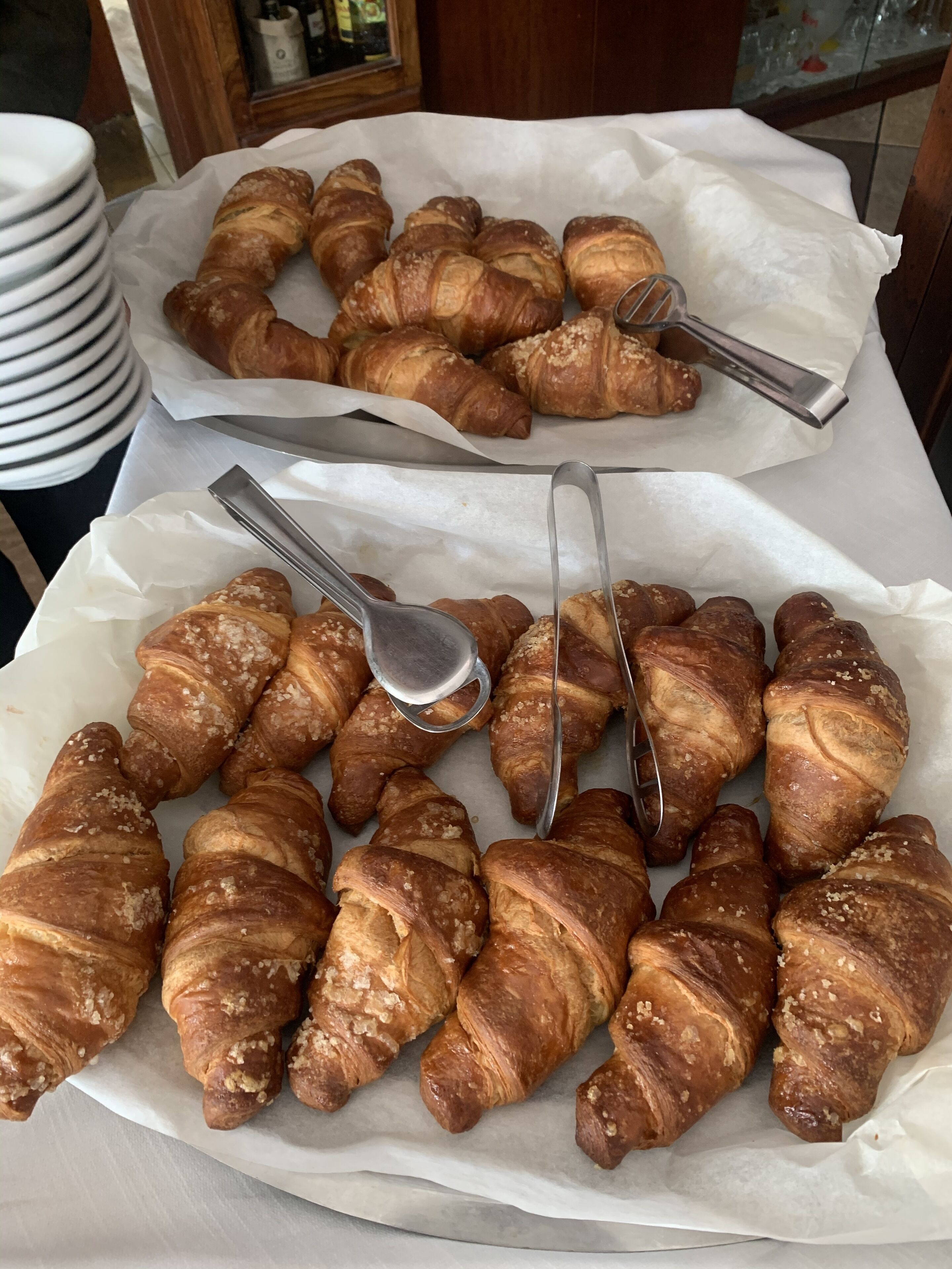 A table with multiple croissants arranged on parchment paper, accompanied by two metal tongs.